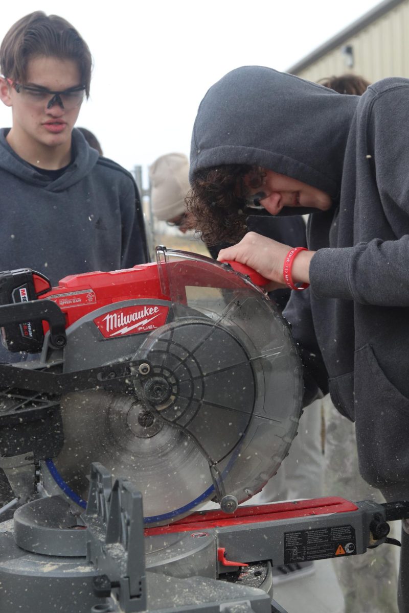 Cutting through. Freshman Maddox Pomeroy watches as freshman Xavier Halbrook uses a miter saw. “I was cutting a 2x4 for a cornhole set,” Halbrook said.
