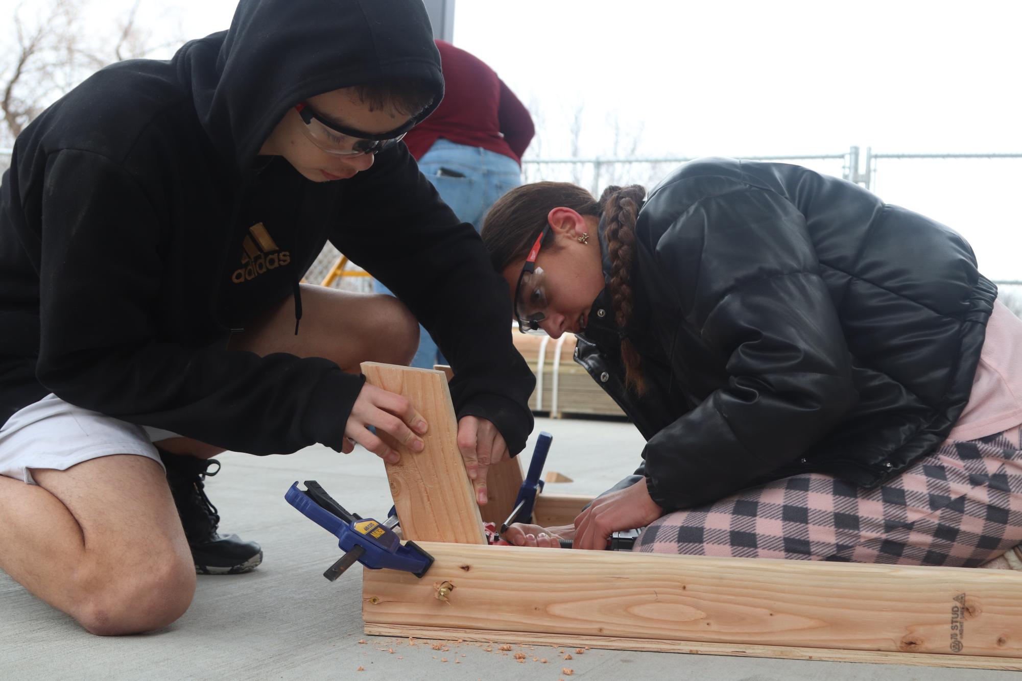 Collaborating. Sophomore Tristen Moore holds wood pieces together while freshman Gracelyn Fikki drills them together. They are building a cornhole set for their upcoming cornhole tournament. “We are drilling a hole through the leg and side of the cornhole board, where the screw will go in order to attach the leg,” Fikki said. 
